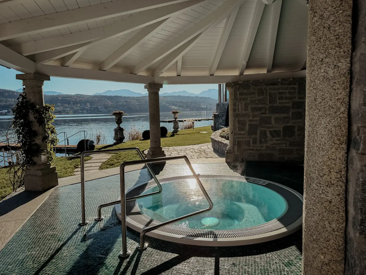 Ein runder Whirlpool im Freien - der SchlossSeefels Spa Whirlpool - ruht unter einem weißen, offenen Pavillon mit Blick auf einen malerischen See und die Berge. Das Sonnenlicht glitzert auf dem Wasser, während Grünpflanzen und eine Steinmauer zu diesem ruhigen COCOtravel-Ausflug beitragen.